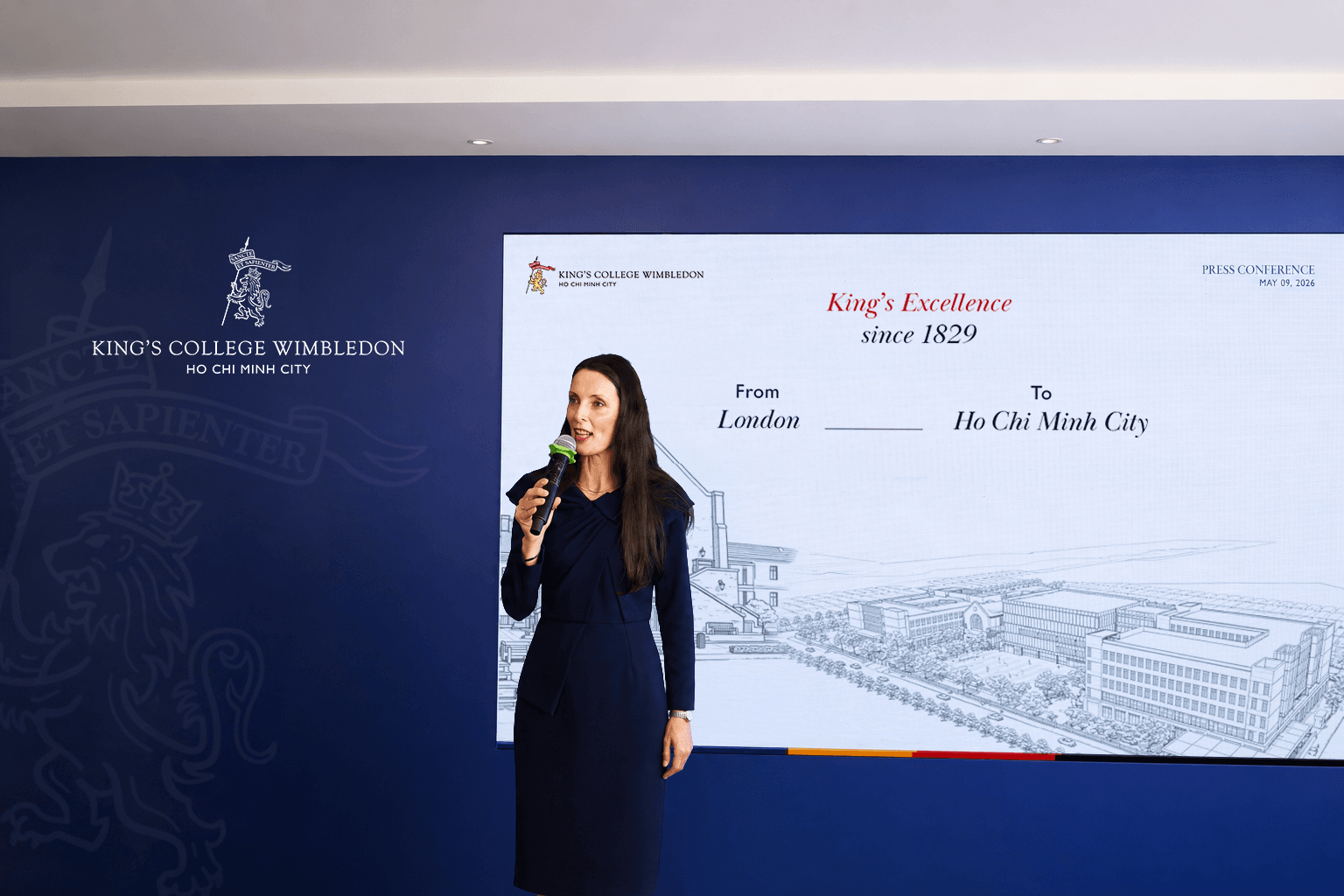Woman speaks into microphone at a King's College Wimbledon event with a screen displaying 'King's Excellence'.