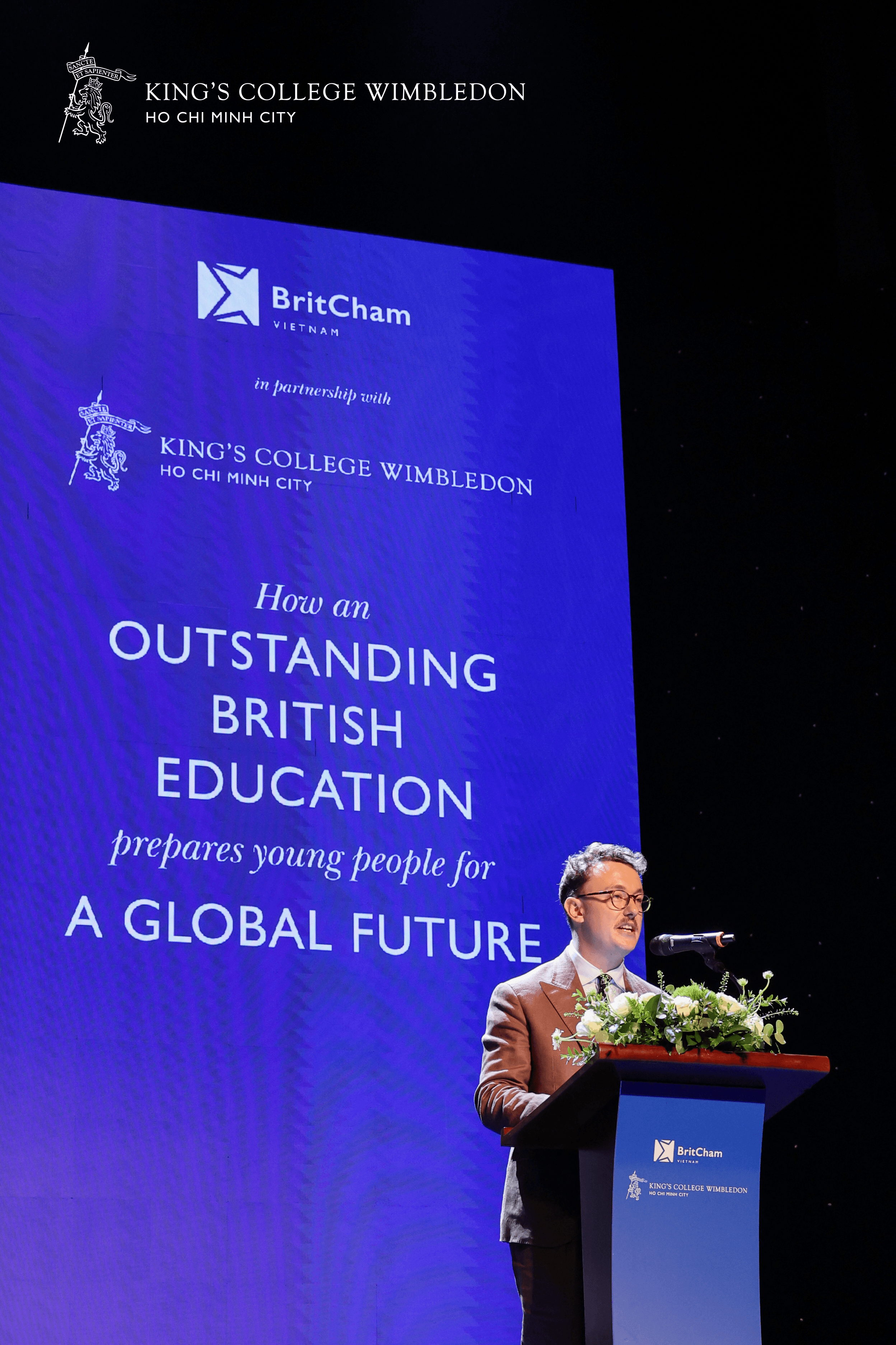 Man speaks at a podium in front of a screen with 'Outstanding British Education' text.