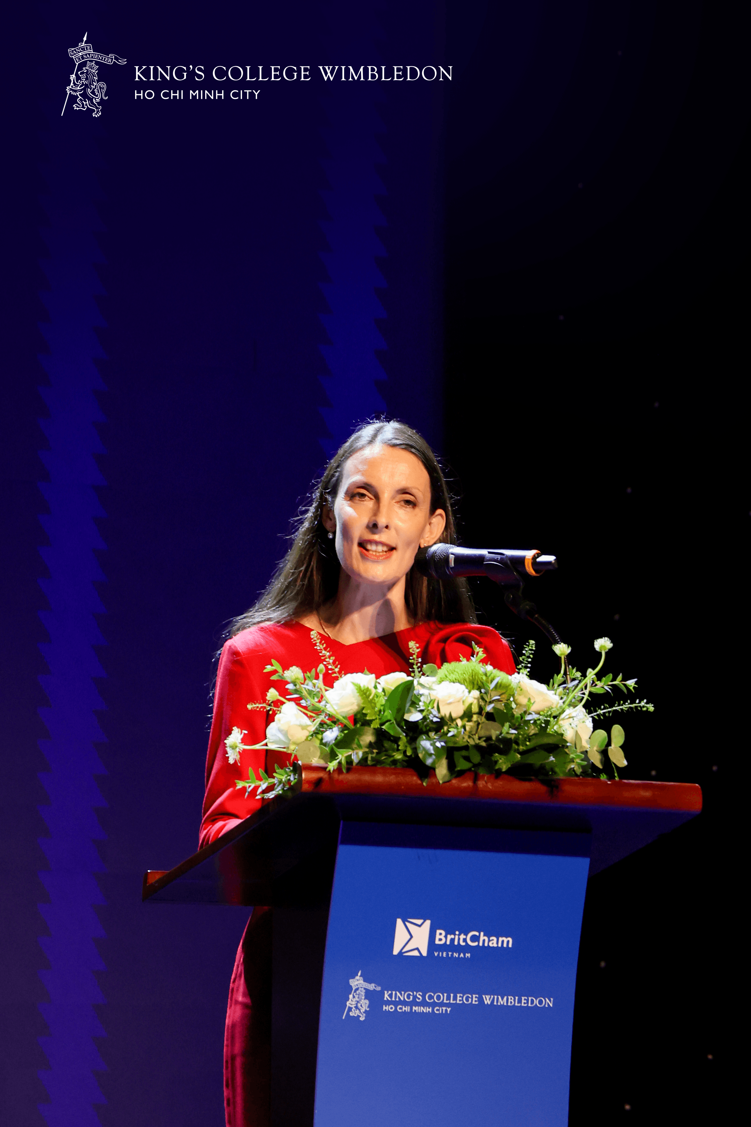 A woman in a red dress speaks at a podium adorned with white flowers.
